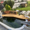 Zeigt einen schönen kleinen Koi Teich im Steingarten-Stil, mit einem interessanten Wassereinlauf sowie einer Art Brücke/Plattform, welche zum verweilen und abschalten einlädt.